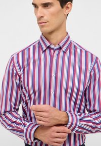 Camicia a maniche lunghe con righe verticali in rosso, blu e bianco. Presenta un colletto button-down e dettagli a contrasto sui polsini. Tessuto dalla superficie liscia.