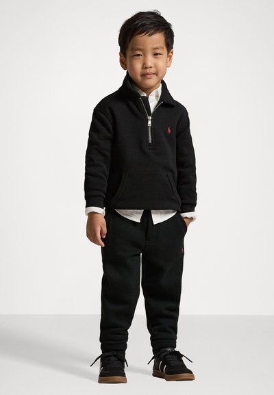 Schwarzer Reißverschluss-Pullover mit einer Fronttasche und rotem Logo, kombiniert mit schwarzen Sweatpants. Das Outfit wird durch ein weißes Hemd mit Kragen darunter ergänzt.