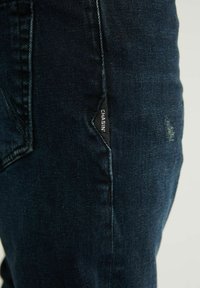 Jean en denim foncé à coupe slim, avec une étiquette noire sur le côté portant la mention 'CHASIN'. Texture lisse avec des détails de décoloration subtile.
