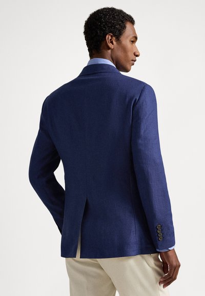 Vyras vilkintis mėlyną švarką ir smėlio spalvos kelnes, stovintis šonu, viena ranka kišenėje, priešais paprastą šviesų foną.