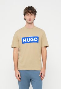 Mladý muž kučeravými vlasmi v béžovom tričku s nápisom "HUGO" tučnými bielymi písmenami na modrom obdĺžniku, doplnenom modrými nohavicami.