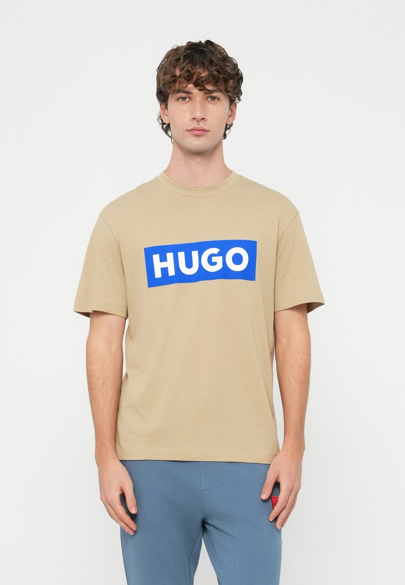 Mladý muž kučeravými vlasmi v béžovom tričku s nápisom "HUGO" tučnými bielymi písmenami na modrom obdĺžniku, doplnenom modrými nohavicami.