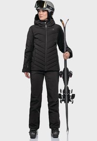 Schwarze isolierte Skijacke mit Chevron-Steppung, kombiniert mit einer passenden schwarzen Skihose. Das Model hält Skier und trägt einen Helm sowie eine Skibrille.