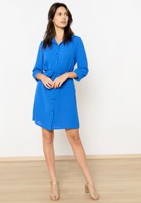 Gaun biru dengan kerah, lengan tiga perempat, pinggang disleting, dan pola titik yang halus. Dipadukan dengan sandal bertumit blok dengan tali.