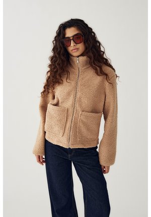 Naine, kellel on pikad lokkis juuksed, kannab üle suure pruunid päikeseprillid, beeži värvi ees lukuga fliisjakka suurte esitaskutega ja tumedaid siniseid teksaseid.