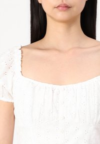 Robe blanche cintrée avec de courtes manches bouffantes, broderie florale texturée, décolleté carré et boutons à l'avant. Tissu doux et léger.