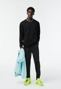 Jonge man die in een zwart outfit staat en een lichtblauwe jas vasthoudt, met neongele sneakers op een witte achtergrond.