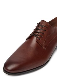 Hnědé kožené společenské boty s špičatou špičkou, hladkým povrchem a tónovanými tkaničkami. Disponují jemným prošíváním a minimalistickými designovými akcenty.