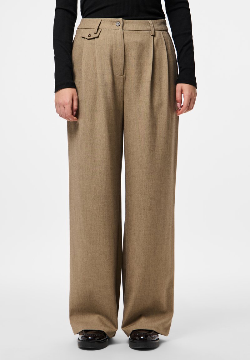 Γυναίκα που φοράει μπεζ παντελόνι με φαρδιά μπατζάκια και ψηλή μέση, συνδυασμένο με μαύρη μακρυμάνικη μπλούζα και μαύρα γυαλιστερά παπούτσια, στέκεται μπροστά σε λευκό φόντο.