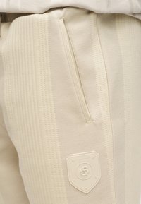 Pantalon beige texturé avec des rayures verticales, doté d'une poche zippée cachée et d'un patch logo en relief en forme de bouclier sur le devant.