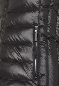 Melnā pūkaina jaka ar quilted dizainu, kurai ir kontrastējoša melna rāvējslēdzēja kabata priekšpusē un gluda, spīdīga tekstūra.