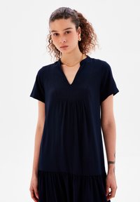 Vestido azul marino con mangas cortas, escote en V, diseño plisado, falda acampanada y un acabado texturado. Acentuado con un collar de cadena dorada.
