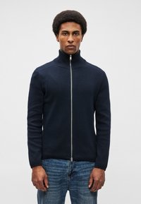 Σκούρο ναυτικό μπλε φούτερ με φερμουάρ και υψηλό γιακά, ριγωτή υφή και ασημένιο φερμουάρ. Φοριέται με μπλε τζην, που διαθέτουν άνετη εφαρμογή.