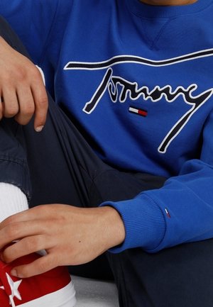 Sweat-shirt bleu royal avec logo "Tommy" brodé en blanc, poignets côtelés, associé à des baskets rouges avec des accents blancs.