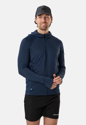 Mann trägt eine marineblaue Kapuzenjacke mit Reißverschluss und Daumenlöchern sowie schwarze Sportshorts, steht mit verschränkten Händen und lächelt leicht.