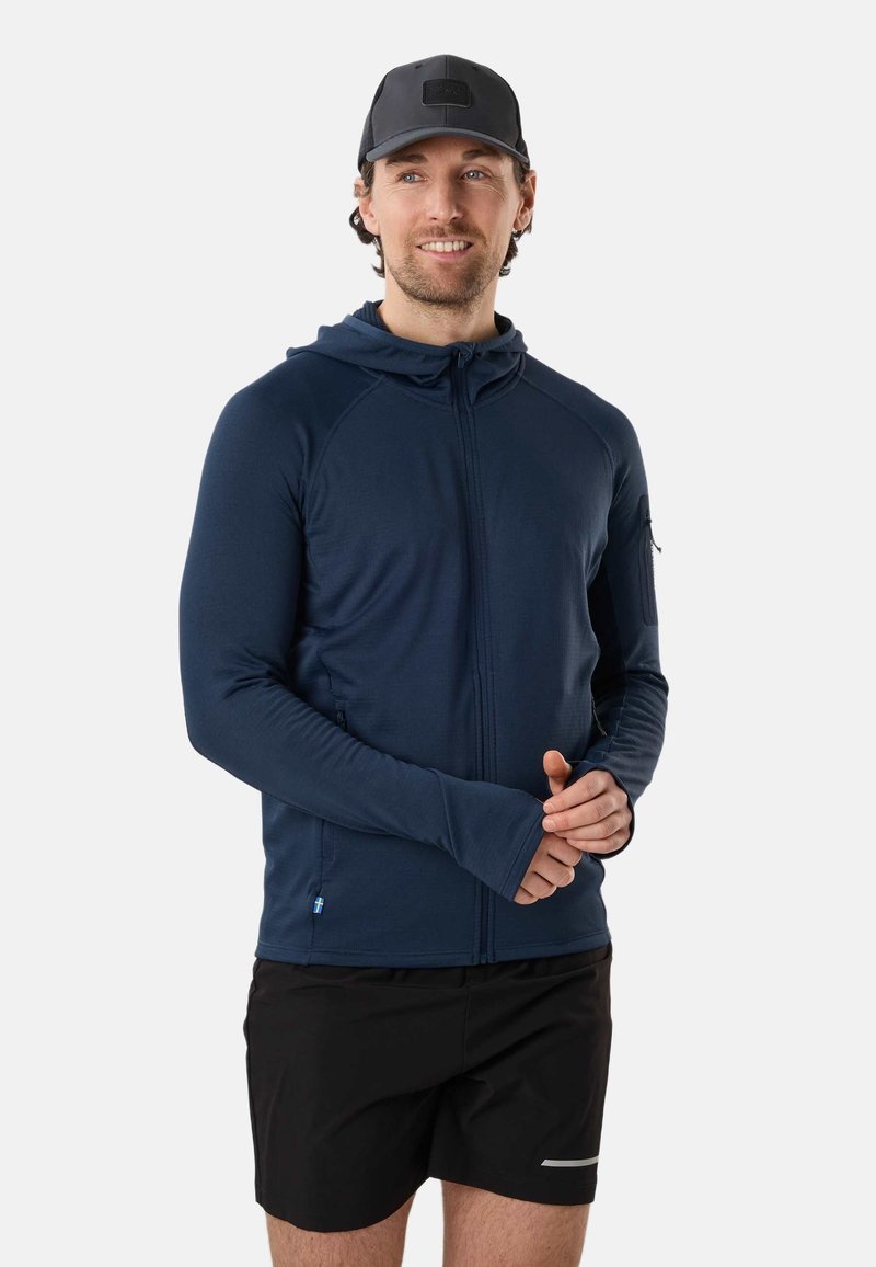 Homme portant un sweat à capuche bleu marine zippé avec des ouvertures pour les pouces et un short de sport noir, debout les mains jointes, souriant légèrement.