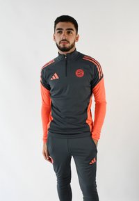 Chándal gris de Adidas con acentos naranjas. Presenta un cierre completo, mangas largas y pantalones ajustados. Logo del Bayern de Múnich en el pecho y el muslo.