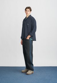 Jūrų mėlynos langelių rašto marškiniai su laisvu sijonu, dėvimi kartu su tamsiomis kelnėmis, turinčiomis mėlyną šoninio juostą. Modelis dėvi šviesiai rudas šlepetes.