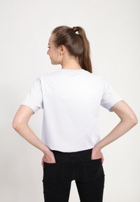 Mladá žena v jednoduchém bílém tričku a černých kalhotách, s rukama v zadních kapsách, otočená zády k neutrálnímu pozadí.