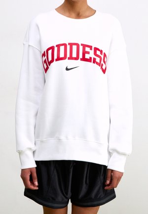 Osoba v bílém svetru s nápisem "GODDESS" červeně a černým logem Nike swoosh, v kombinaci s černými sportovními šortkami.