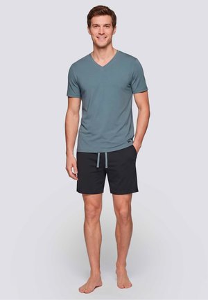 Lachende man staat blootsvoets, gekleed in een blauw T-shirt met korte mouwen en V-hals en zwarte korte broek met trekkoord, tegen een effen lichte achtergrond.