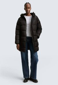 Sort dunjakke med hætte, der har quiltede sektioner og en lynlås foran. Kombineret med blå vidde bukser og sorte støvler.