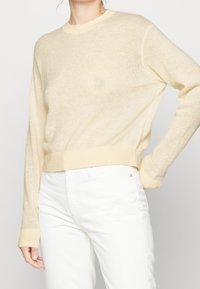 Femme portant un pull en maille beige clair à manches longues et un jean blanc taille haute, avec une main sur la hanche et l'autre détendue.
