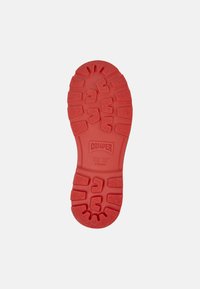 Red shoe sole with a textured pattern and visible brand name "Camper." The design features deep grooves for grip and stability.