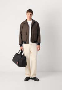 Brown woolen blend jacket met twee grote voorkeurszakken, een wit t-shirt eronder, crèmekleurige wijde broek en een zwarte sporttas. Zwarte schoenen.