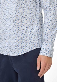 Camicia a maniche lunghe con un motivo floreale azzurro su tessuto bianco, con orlo curvo e dettagli con bottoni. Pantaloni blu scuro parzialmente visibili.