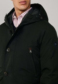 Muškarac nosi crni kaput s kapuljačom preko ružičaste i bijele prugaste košulje i tamnoplave vune.