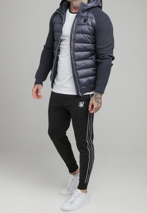 Mand iført navyblå dunjakke med strikkede ærmer, hvid t-shirt, sorte joggers med hvide striber og hvide sneakers, stående på ensfarvet baggrund.