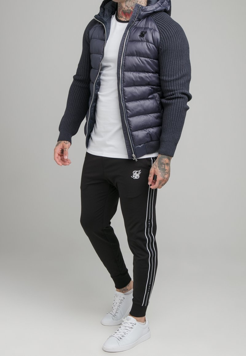 Homme portant une doudoune bleu marine avec des manches en tricot, un t-shirt blanc, un pantalon de jogging noir avec des bandes blanches, et des baskets blanches, debout sur un fond uni.