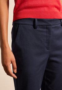 Granatowe spodnie bawełniane z mieszanki, z diagonalną teksturą, bocznymi kieszeniami i szlufkami na pasek. Ręka opiera się na spodniach, ukazując zbliżenie na pas.