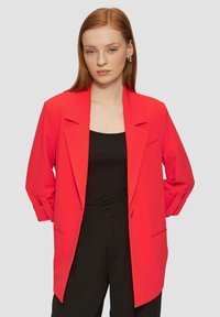 Blazer rouge au design ajusté, épaules structurelles et silhouette longue. Il dispose d'un bouton unique et de poignets retroussés, porté sur un haut noir.