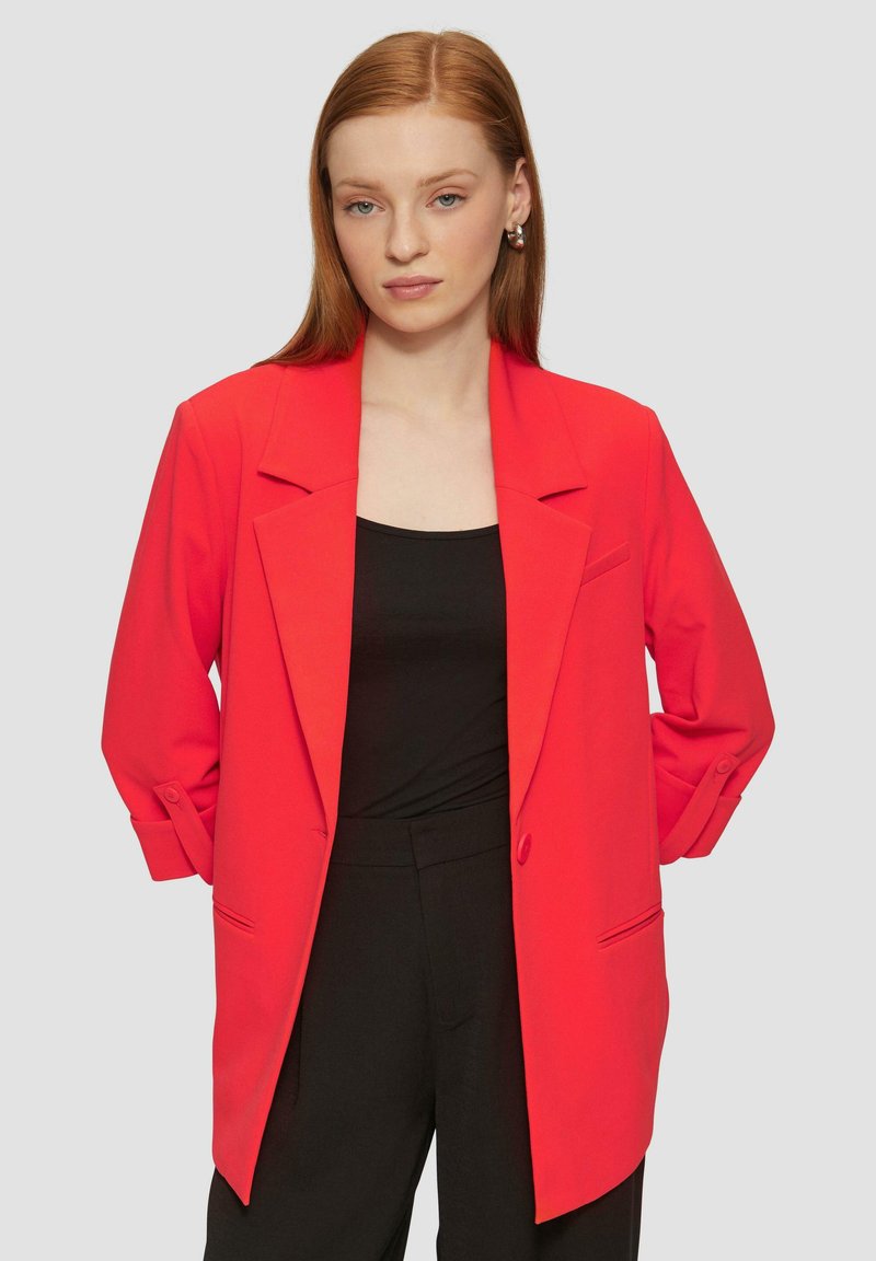 Blazer rouge au design ajusté, épaules structurelles et silhouette longue. Il dispose d'un bouton unique et de poignets retroussés, porté sur un haut noir.