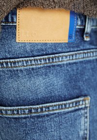 Blå denimjeans har ett strukturerat tyg, med en läderlapp på midjebandet som visar präglande text och en blå accent.
