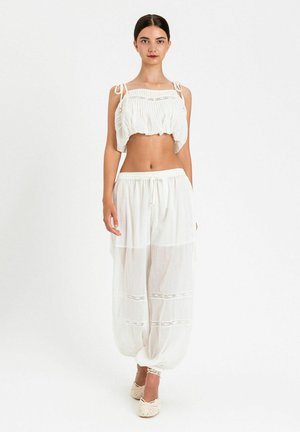 Donna in piedi che indossa un top corto bianco senza maniche con lacci e pantaloni bianchi larghi con dettagli in pizzo e scarpe annodate alla caviglia su sfondo bianco.