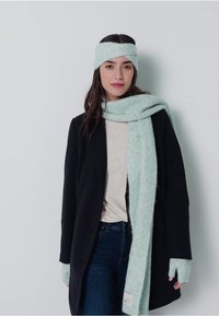 Ljusgrön stickad scarf, matchande pannband och fingerlösa handskar som bärs med en svart kappa och beige topp. Mjuk textur, ribbat mönster.