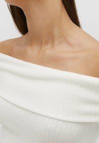 Biała bluzka typu off-shoulder z prążkowanego materiału o gładkiej fakturze, posiadająca szeroki dekolt i miękki, lekki materiał. Zbliżenie na dekolt.