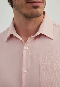 Chemise rose clair à manches courtes avec un motif texturé, un col pointu et une poche poitrine avec un détail de bouton blanc unique.
