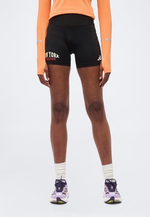 Jambes portant un short noir avec le texte "New York Together", un haut orange à manches longues, des chaussettes blanches et des chaussures de sport violettes sur fond gris.