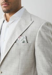 Helles graues kariertes Blazer aus strukturiertem Stoff, mit einer Brusttasche, in die ein gemustertes Taschentuch gesteckt ist. Darunter ein weißes Hemd.