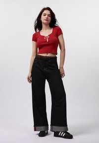 Červený žebrovaný crop top s bílým lemem, spárovaný s širokými černými džíny s ohrnutými lemy a barevným tkaným pásem. Černé boty s bílými pruhy.
