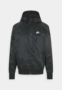 Černá větrovka Nike, která má zapínání na zip, nastavitelnou kapuci, elastické manžety a logo na levé hrudi. Hladký, lesklý materiál.