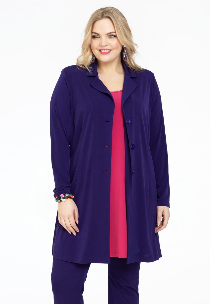 Lang lilla jakke med krave, frontknapper og lange ærmer, båret over en lyserød top. Bemærkelsesværdigt farverigt armbånd på håndleddet. Glat tekstur.