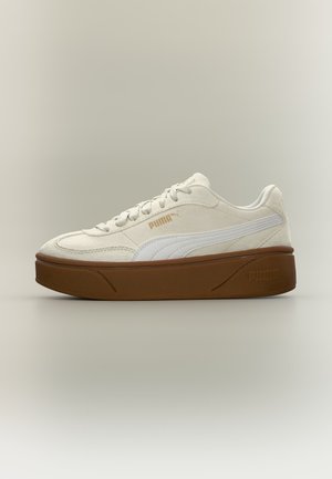 Baskets blanches Puma avec semelle épaisse marron, bande de logo blanche et marquage doré Puma sur le côté, montrées en vue de profil sur fond neutre.