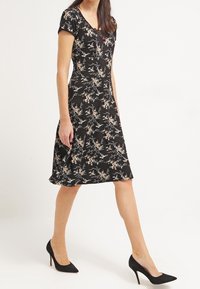 Svart blommig klänning med korta ärmar, V-halsringning och figursydd överdel. Går ner till knäet med en utsvängd kjol. Bärs med svarta högklackade skor.