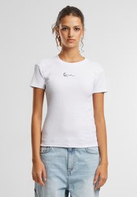 T-shirt en coton blanc à manches courtes, col rond, avec un petit logo noir sur la poitrine. Porté avec un jean en denim bleu clair.
