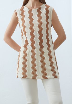 Beżowy i brązowy sweter bez rękawów w zygzakowaty wzór, noszony z białymi spodniami, modelka stoi z rękami za plecami na jednolitym tle.